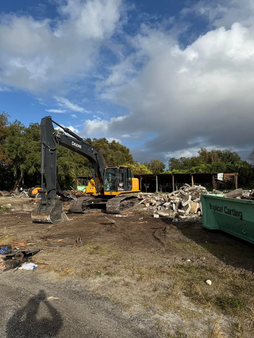 Excavator on construction site clearing debris under a blue sky with clouds.