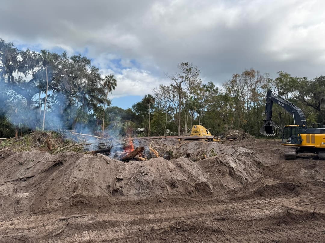 Construction site with burning debris and heavy machinery amid cleared trees and cloudy sky.
