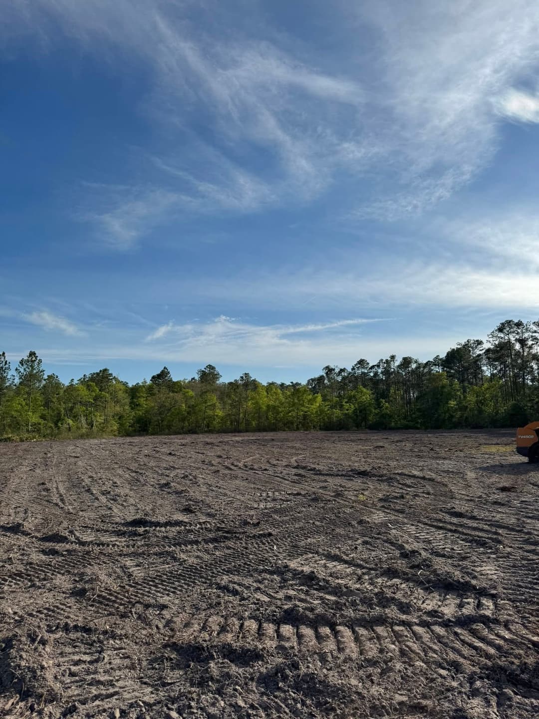 Cleared land with tire tracks under a blue sky and green trees in the background.